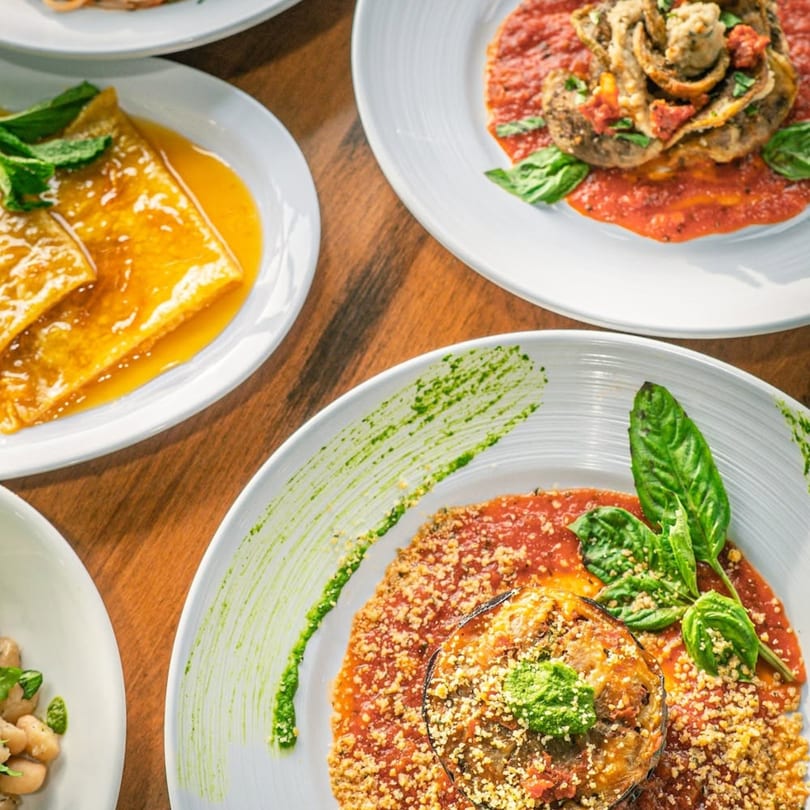 Overhead view of various Italian dishes, including eggplant parmesan, crepes, and pasta, on a wooden table.